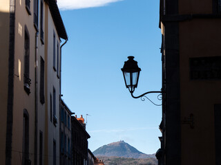 View on a street in Clermont-Ferrand. A street lamp is hung on the wall in the foreground. A mountain is in the background. The city is located in the center of France, in the Auvergne region.