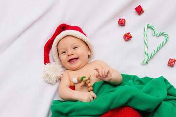 Happy baby wearing handmade crocheted red christmas elf hat and pants, smiling on white fleece blanket with sensory wooden teething toy with crocheted beads