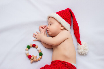 Baby wearing handmade crocheted red christmas elf hat and pants, sleeping on white fleece blanket with sensory wooden teething toy with crocheted beads
