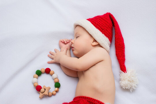 Baby wearing handmade crocheted red christmas elf hat and pants, sleeping on white fleece blanket with sensory wooden teething toy with crocheted beads