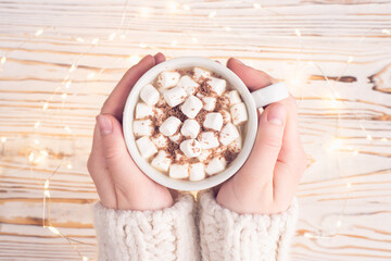 Home atmosphere concept. Top above overhead view photo of female hands holding cup with tasty chocolate and marshmallow over wooden white rustic background with lights