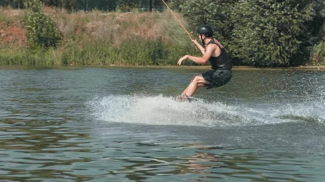 Tracking full shot of athletic boy in water sports clothing standing on wakeboard sliding on lake holding handle splashing water