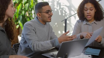 Medium shot of young diverse colleagues sitting at desk with laptop arguing emotionally while discussing business project looking through papers