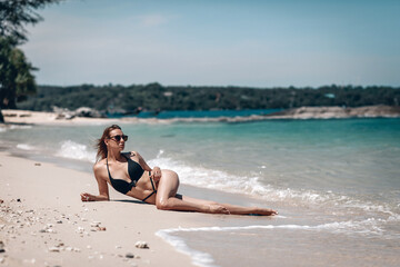 Fototapeta na wymiar Young girl with a gorgeous body is resting on the beach with white sand near the ocean. Beautiful sexy model in a black bathing suit and black sunglasses sunbathing.