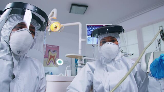Dental team leaning over patient to work on patient gums dressed in hazmat suit in dental office with new normal. Stomatology team wearing safety equipment against coronavirus treating teeth problem.