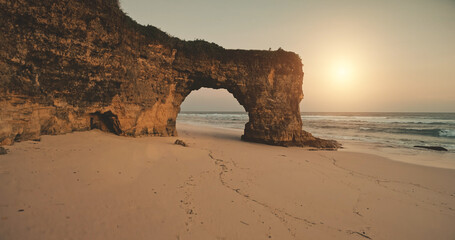 Cliff unique wall with giant hole at sun light aeial. Sand beach at ocean bay waves. Nobody nature scenic seascape. Rock shore of Sumba Island, Indonesia. Cinematic tropic paradise resort