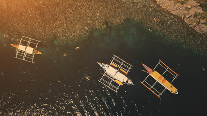 Sunset top down at ocean bay: passengers swim and dive at boats in aerial shot. Tourists leisure at summer cruise tour on traditional vessels. Philippines vacation at tropical island of Palawan