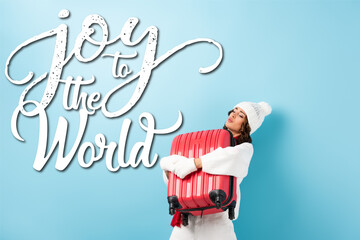  woman in winter outfit carrying heavy suitcase near joy to the world lettering on blue