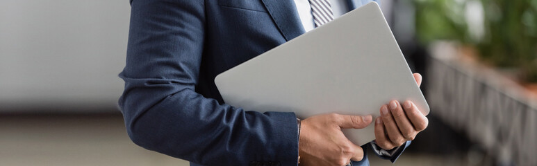 Cropped view of businessman in formal wear holding laptop on blurred background, 