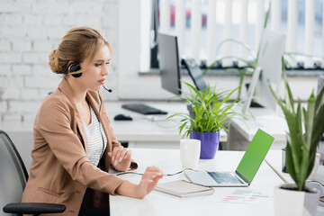  woman in headset looking at laptop, while sitting at workplace with digital devices on blurred...