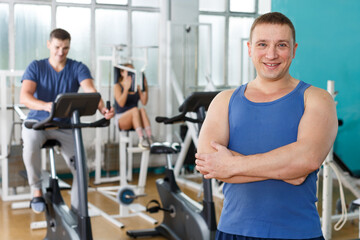 Fototapeta na wymiar Portrait of cheerful sporty man posing at gym with his friends on background