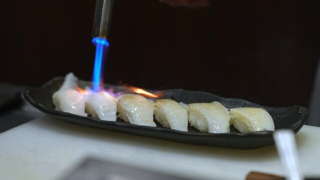 Close up of Professional chef hand use butane torch burner burning engawa sushi on serving plate. Chef preparing healthy menu fish sushi on open kitchen counter bar to customer in japanese restaurant