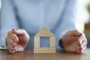 Woman covering wooden house with her hand closeup. Real estate insurance concept