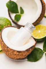Bottle of lotion in coconut half near lime, and rose leaves on blurred white