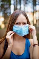 Beautiful young brunette woman with long hair wearing medical mask in a pines and firs forest on a summer day during the covid-19 pandemic.