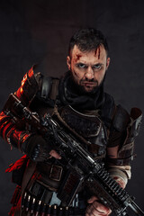 Dressed in dirty clothing with dark armour bearded survivor holding shotgun poses in dark background with serious face.