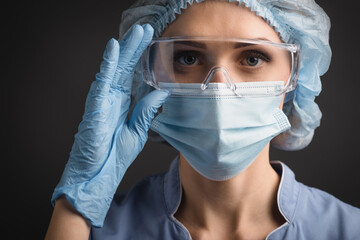 nurse in blue medical cap and mask adjusting goggles while looking at camera isolated on dark grey, stock image