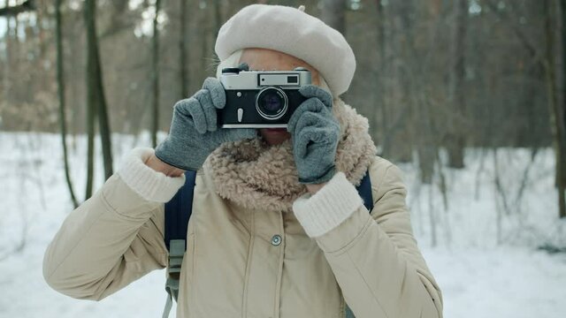 Portrait of senior woman photographer taking picture with modern camera smiling and looking at camera in winter forest. Photography and people concept.