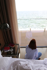 
a girl sits on the floor by the bed and looks at the sea