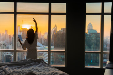 Fototapeta na wymiar Asian woman practicing yoga, exercise near bed with beautiful sunlight in the morning and city scenery in the window. Use tablet device listen music, application and reading message. Copy space