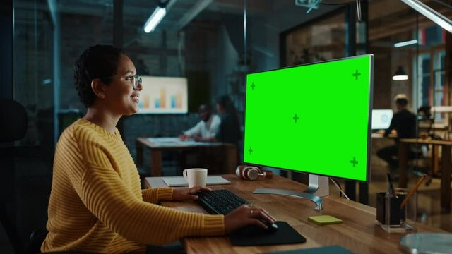 Young Multiethnic Specialist Working on Desktop Computer with Green Screen Mock Up Display in Creative Office. Beautiful Diverse Female Manager with Short Hair and Glasses is Wearing a Yellow Jumper.