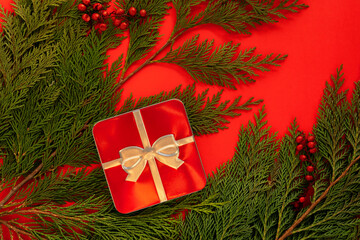 Red metal box with silver bow on top on bright red background with juniper branches around as a Christmas gift concept, celebration, festive time. Creative present concept.