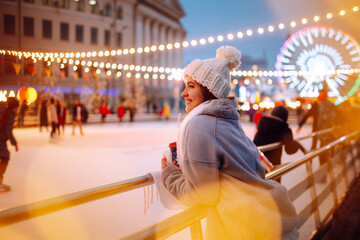 Smiling woman in winter style clothes with coffee near skating rink. Young woman enjoying winter holidays on Christmas market. Lights around.