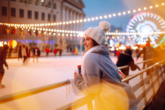 Smiling woman in winter style clothes with coffee near skating rink. Young woman enjoying winter holidays on Christmas market. Lights around.