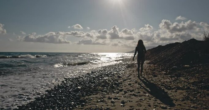 Silhouette of attractive girl walking along shore. Slender and sexy girl moving on sandy beach. The seascape and the glare of the sun. Young woman cinematic strolling. Concept of nature and beauty