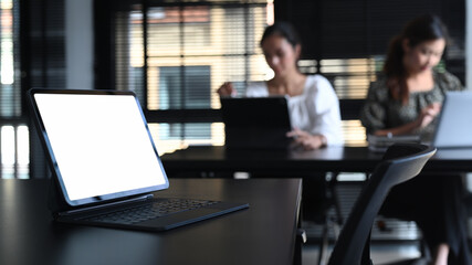 A blank screen computer tablet on black wooden table with two businesswoman sitting in office blurred background.