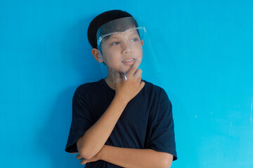 Asian boy used face shield standing over blue background looking confident and smiling with crossed arms and hand raised on chin. Thinking positive.