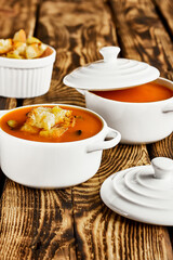 Vegetarian spicy autumn pumpkin cream soup served in white cocotte with croutons and pumpkin seeds on wooden table. Seasonal autumn vegetarian food.