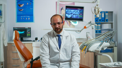 Man stomatologist speaking on video camera sitting on chair in dental clinic with assistent in background. Dentistry doctor looking on webcam explaining treatment while nurse is working on computer