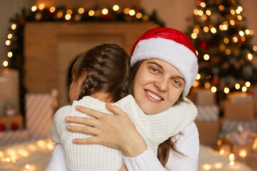 Mom and daughter near Christmas tree and fireplace hugging each other with happiness, wearing white shirts and santa hat, happy holidays at home, congratulating with New Year.