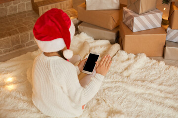 Little girl holding phone with blank screen while posing backwards, wearing white sweater and santa hat, sitting on floor on soft carpet. Merry Christmas and Happy Holidays!