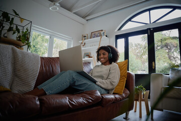 Happy girl wearing headphones watching and listening videos on laptop sitting on couch at home