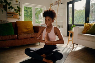 Young african woman sitting in lotus position with joined hands on yoga mat practicing breathing...