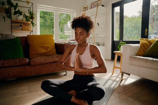 Young african woman sitting in lotus position with joined hands on yoga mat practicing breathing exercise