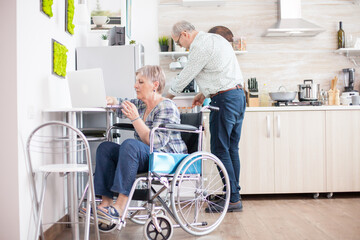 Handicapped woman typing on laptop in the kitchen. Disabled old woman in wheelchair working. Paralyzed handicapped old elderly person using modern communication online internet web techonolgy.