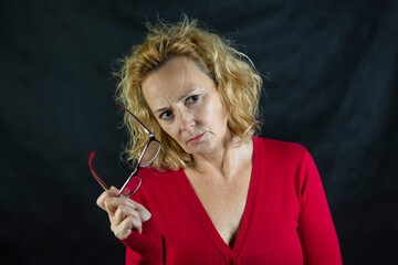 Obraz premium Middle-aged blonde woman looks with an angry expression holding up her glasses dressed in a red sweater on a dark background