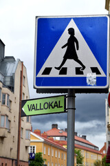 Under pedestrian crossing sign, is a sign in Swedish informering where the nearby polling station is located on election day. Buildings in the background.