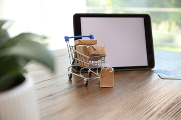 Internet shopping. Small cart with boxes and bag near modern tablet on wooden table indoors