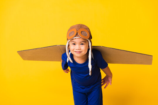 Happy Asian Handsome Funny Child Or Kid Little Boy Smile Wear Pilot Hat Playing And Goggles With Toy Cardboard Airplane Wings Flying, Studio Shot Isolated Yellow Background, Startup Freedom Concept