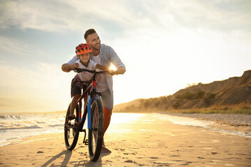 Happy father teaching son to ride bicycle on sandy beach near sea at sunset