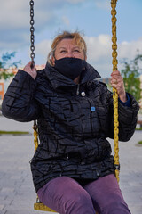 A 68-year-old woman wearing a black medical mask sits on a swing in the town square