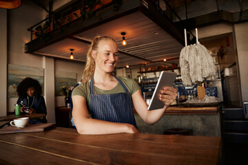 Smiling female waiter standing leaning on table using digital tablet in modern cafe