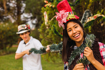 Young couple having fun while decorating the Christmas tree - Beautiful young brunette woman in a Christmas hat smiling and the young brunette holding a garland.