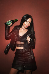 Sexy punk brunette woman in leather jacket leather skirt and lace bralette posing with beer red wall background hard light. Film toned grain add image.