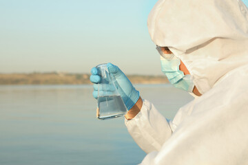 Scientist in chemical protective suit with conical flask taking sample from river for analysis