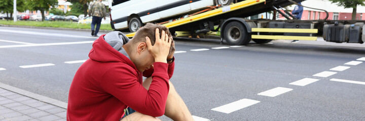 Minivan driver sits on side of road and holds his head during evacuation of his vehicle. Tow truck service business concept.